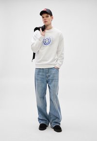 Λευκό φούτερ με μπλε γραφικό χαμογελαστό πρόσωπο και κείμενο, συνδυασμένο με φαρδιά ανοιχτό μπλε τζιν παντελόνια και μαύρα παπούτσια slip-on. Μαύρο καπέλο φορεμένο ανάποδα.