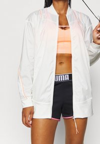 Valkoinen urheilujakku oransseilla yksityiskohdilla, vetoketjullinen etuosa, yhdistetty persikanväriseen crop toppiin ja mustiin shortseihin, joissa on vaaleanpunainen viimeistely ja logovyötärö.