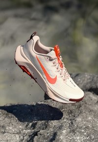 Nike running shoe in light beige and red. Mesh upper, cushioned sole, lightweight design, visible branding, and textured accents.