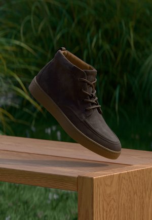 Bota de ante marrón hasta el tobillo con suela de goma y cordones suspendida sobre un banco de madera al aire libre con plantas verdes desenfocadas en el fondo.