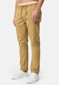 Pantalones khaki con cintura elástica y cordón, bolsillos laterales y corte ajustado. Combinados con zapatillas blancas sobre un fondo sencillo.