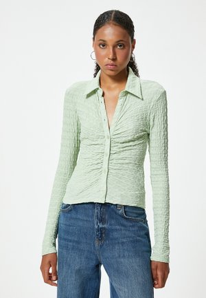 Blouse longue à manches longues de couleur vert clair et texturée avec un devant à boutons et un col pointu, associée à un jean en denim bleu.