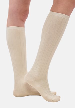 Jalad, millel on põlveni ulatuvad beežid soonilised sokid, üks jalg toetub varvastele, taustaks tavaline valge taust.