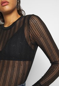 Haut long à manches en mailles noires avec des rayures verticales et un éclat subtil. Doté d'un col rond et d'une coupe ajustée, superposé sur un bralette triangulaire.