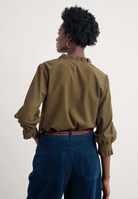 Alyvuogių žalumo viršūnė su voliotais apykaklėmis ir kamuoliais rankovėmis, derinama su mėlynomis korduroy kelnėmis ir ruduoja audiniu diržu. Rodomas galinis vaizdas.