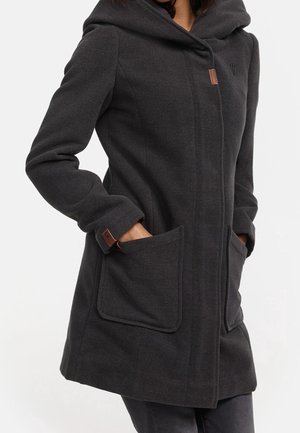 Femme portant un manteau à capuche gris foncé avec de grandes poches avant, les mains partiellement dans les poches, et une fermeture avant dissimulée.