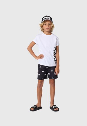 Blondes Kind steht mit einer Hand in der Hüfte, trägt ein weißes T-Shirt, schwarze Shorts mit weißem Muster, schwarze Mütze und schwarze Slides mit dem Schriftzug "KARL".