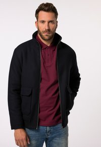 Navyblaue Jacke mit Reißverschluss und hohem Kragen. Verfügt über zwei Fronttaschen und elastische Bündchen. Wird über einem rotbraunen Poloshirt und blauen Jeans getragen.
