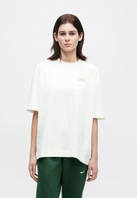 Άτομο με ίσια σκούρα μαλλιά φοράει λευκό oversized T-shirt με μικρό χρυσό κείμενο, σε συνδυασμό με σκούρα πράσινα Nike παντελόνια φόρμας, στέκεται μπροστά σε άσπρο φόντο.