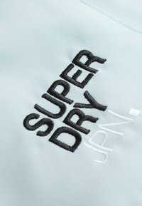 Gaiši zila auduma virsma ar izšūtu melnu tekstu "SUPER DRY" un baltajiem akcentiem. Gluda tekstūra, tuvplāns ar šuves detaļām.