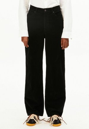 Personne portant un pantalon noir à jambes larges, une chemise blanche à manches longues et des baskets noires avec des lacets blancs épais et des semelles orange, debout.