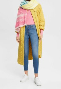 Abrigo acolchado mostaza, suéter de punto rosa, jeans ajustados azules y zapatillas blancas. Bufanda multicolor con tonos rosa y azul, con flecos.