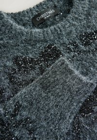 Pull gris foncé duveteux avec texte noir en sequins et une étiquette noire indiquant « CHRISTMAS AT NEXT » au niveau de l'encolure.