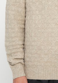 Beige pletený svetr se svrškem a rukávy s texturovaným geometrickým vzorem, žebrovanými manžetami a rukou odpočívající vedle těla.