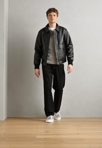 Veste bomber en cuir noir avec deux poches avant, pull gris en dessous, pantalon noir ample et baskets blanches. Fond en parquet clair.