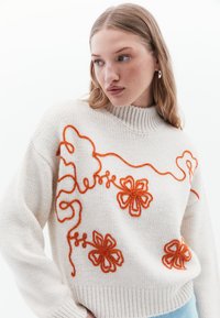 Maglione lavorato a maglia crema con vivace ricamo floreale arancione e motivi a spirale. Il collo e i polsini a coste esaltano la texture.
