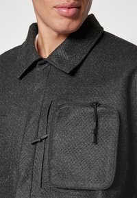 Veste noire texturée avec col, dotée d'une poche zippée et de détails perforés à l'avant pour un design amélioré.
