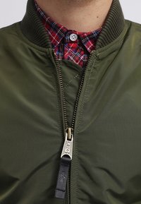 Ingrandimento di una giacca bomber verde oliva parzialmente zipata sopra il colletto di una camicia a quadri rossa e blu abbottonata.