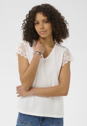 Femme aux cheveux bouclés portant un haut blanc à manches en dentelle et un jean bleu, tenant son bras gauche avec sa main droite devant un fond uni.