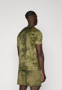 Olijfgroene sportset bestaande uit een shirt en shorts, met een gestructureerd, patroonoppervlak met een camouflageontwerp. Korte mouwen en een ronde hals.