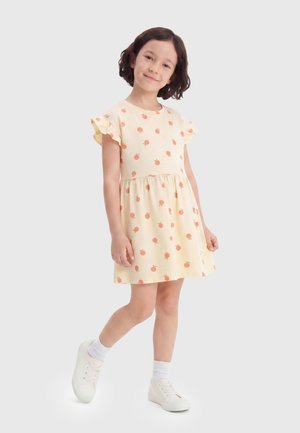 Giovane ragazza sorridente, indossa un vestito giallo con stampa di frutta arancione, calzini bianchi e scarpe da ginnastica bianche, in piedi con una gamba leggermente sollevata.