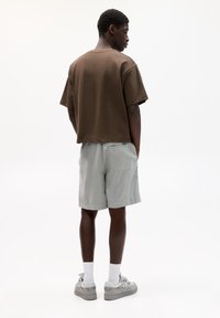 Smeđa pamučna košulja kratkih rukava uparena sa svijetlosivim šortsevima. Outfit se odlikuje opuštenim krojem i minimalističkim dizajnom. Ležerna obuća upotpunjuje izgled.