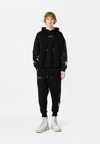 Sudadera negra con detalles de flecos en las mangas, combinada con joggers negros que tienen un bolsillo con cremallera. Llevada con botas de combate blancas.