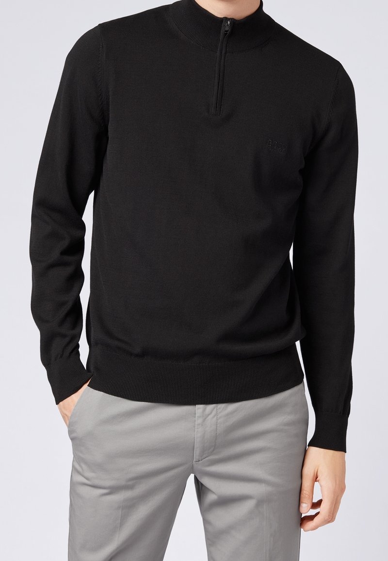 Muž v černém dlouhém zipovém svetru a světle šedých kalhotách, s jednou rukou v kapse a viditelným logem značky na svetru.