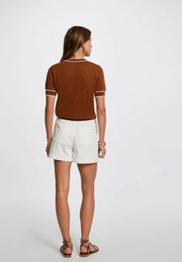 Top corto a maniche corte in maglia marrone con bordi bianchi abbinato a pantaloncini bianchi. I pantaloncini hanno tasche posteriori e un design aderente. Sandali marroni con borchie.