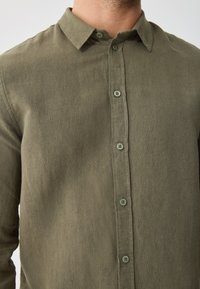 Olivově zelená košile na knoflíky z texturované látky. Má klasický límec a šest knoflíků stejné barvy po celé přední části.