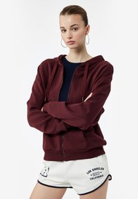 Sudadera burdeos con cremallera y capucha, de textura suave y mangas largas, llevada sobre una camiseta negra. Combinada con pantalones cortos blancos con acentos en azul marino.