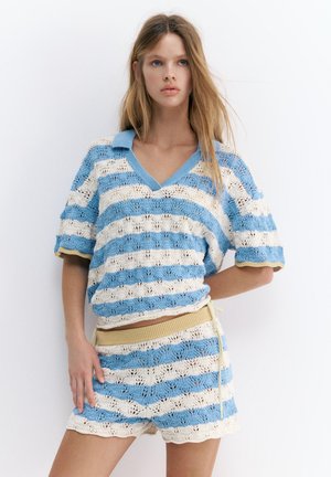 Młoda kobieta w niebiesko-białym pasiastym dzianinowym topie z kołnierzykiem i pasujących szortach, stojąca na białym tle z ręką na biodrze.
