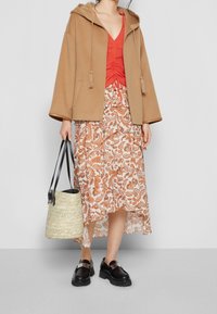 Cappotto beige con cappuccio, top rosso strutturato, gonna a strati stampata in bianco e arancione, mocassini neri e una borsa intrecciata con manici neri.