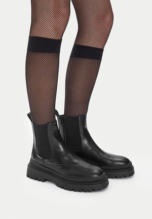 Piernas con medias de red negras combinadas con botas negras gruesas de cuero hasta el tobillo con paneles laterales elásticos.