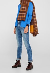 Cardigan blu a maniche lunghe con bottoni, jeans azzurri, e stivaletti alla caviglia in pelle verniciata rossa scura. Sciarpa a quadri marrone e gialla drappeggiata.