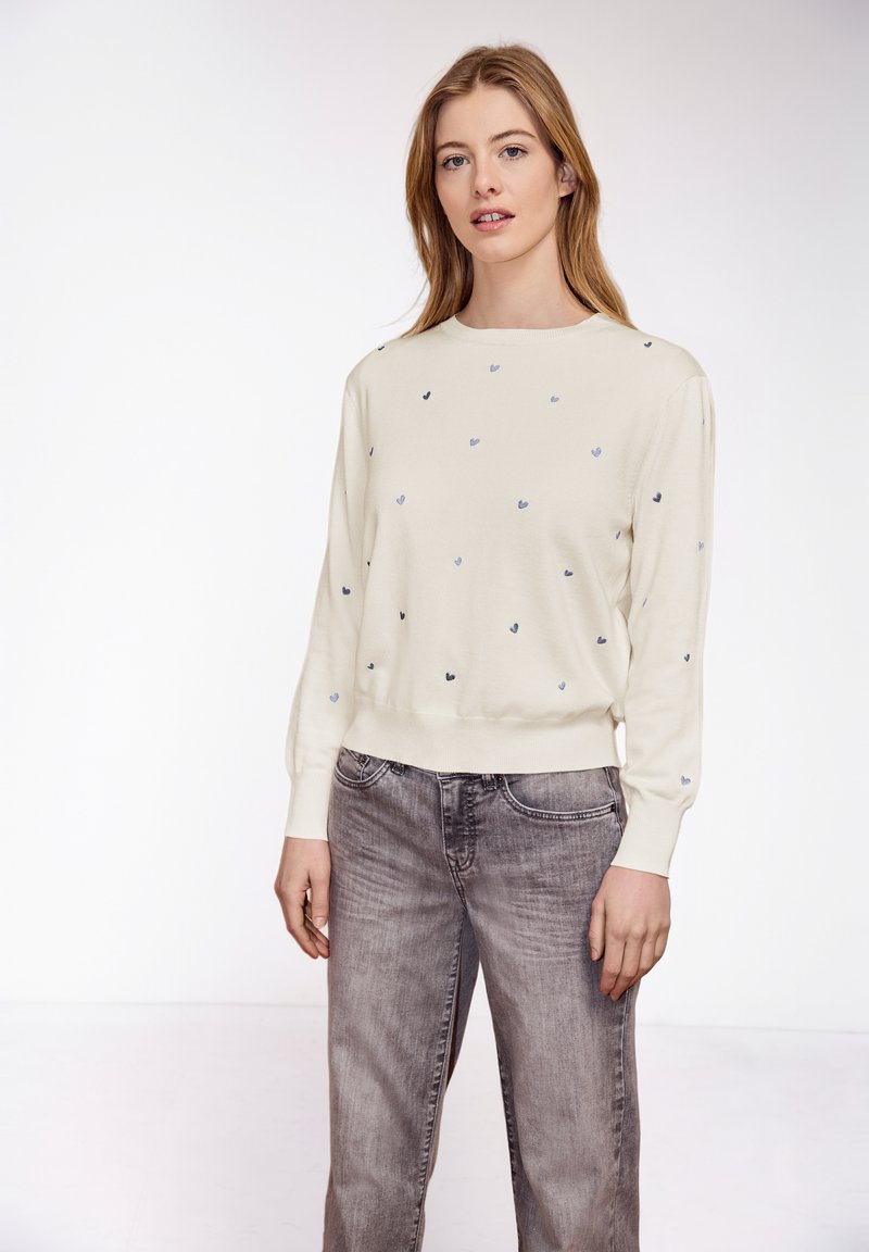 Femme portant un pull crème avec de petits motifs de cœurs, associée à un jean gris délavé, debout devant un fond blanc uni.