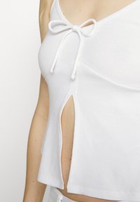 Top de alças branco com textura em ribas, detalhado com um laço no decote e uma fenda nas laterais, apresentando alças finas e um design justo.