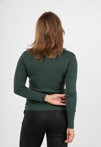 Camicia a maniche lunghe, di colore verde scuro, realizzata in tessuto elastico, design aderente, con una texture liscia e senza motivi visibili. Abbinata a leggings neri e lucidi.