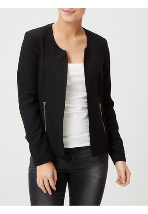 Veste noire courte avec un col rond, en tissu texturé, et des accents de fermeture éclair argentée sur les poches avant. Portée par-dessus un haut blanc.