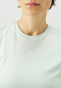 Šviesiai mėtų žalios spalvos sportinė palaidinė su apvaliu kaklu ir trumpomis rankovėmis. Pirmame plane – tekstūruotas baltas logotipas, esantis krūtinės srityje. Glotni medžiaga.