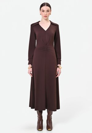 Femme portant une robe marron foncé à manches longues avec un col en V et des bottes marron assorties sur un fond clair uni.