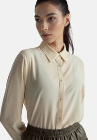 Jasnobeżowa koszula na guziki z długimi rękawami, klasycznym kołnierzykiem i drobnymi guzikami, wykonana z gładkiej tkaniny.