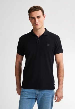Camisa polo negra de manga corta, con una tapeta de tres botones y un parche cuadrado con el logo en el pecho. Se lleva con unos vaqueros de color azul claro.