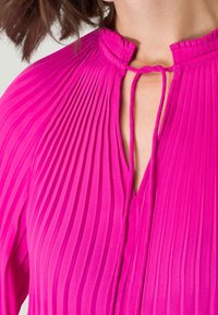 Blouse rose vif avec des plis verticaux, un col montant et une ouverture en forme de clé, ainsi que des liens au cou pour un ajustement réglable.