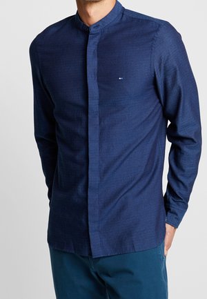 Homme portant une chemise bleu marine à manches longues avec col mandarin, boutons cachés et petit logo sur la poitrine, associée à un pantalon bleu.