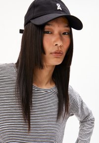 Schwarze Baseballkappe mit weißem Logo, kombiniert mit einem langärmeligen Shirt in schwarz-weißen Streifen. Glatter Stoff und lässige Passform.