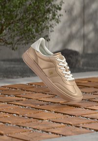 Αθλητικό παπούτσι σε ανοιχτό μπεζ χρώμα με λείο δέρμα, λευκές λεπτομέρειες, επίπεδα κορδόνια και λεπτή σόλα από καουτσούκ. Φέρει διακριτικό λογότυπο.