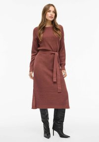 Strikkekjole i rustfarge med teksturert mønster, lange ermer, belte i midjen og en splitt som strekker seg til knærne. Kombinert med svarte støvler som går til knærne.