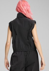 Gilet sans manches noir avec un col haut, en tissu texturé, taille élastique et coupe ample, montrant un tatouage partiellement visible sur le bras du porteur.