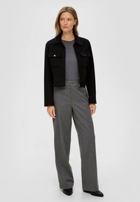 Vrouw staat in een zwarte cropped jas, een grijs langemouwen shirt, grijze high-waisted wijde broek en zwarte puntige schoenen.